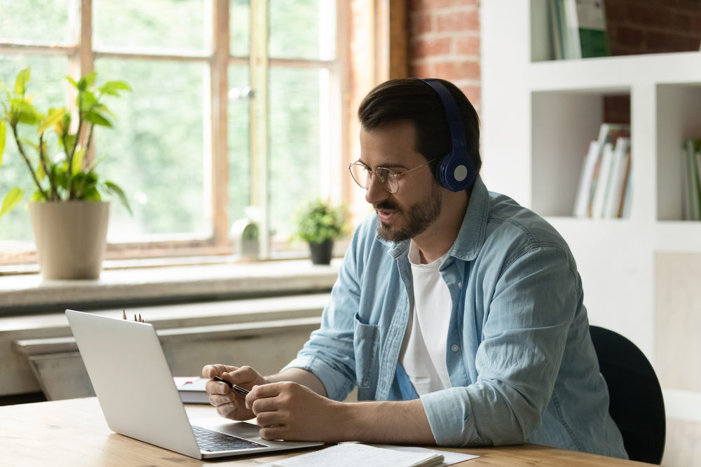 Young Caucasian businessman in earphones work on computer online talk speak on video call with client. Male employee in headphones use laptop have webcam digital conference with customer