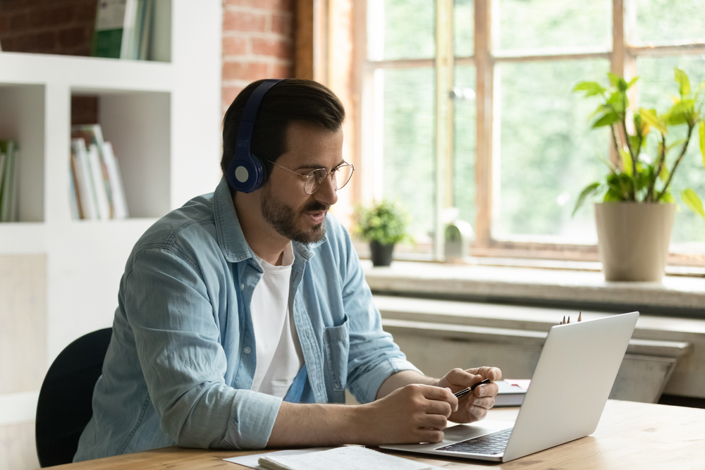 Young Caucasian businessman in earphones work on computer online talk speak on video call with client. Male employee in headphones use laptop have webcam digital conference with customer
