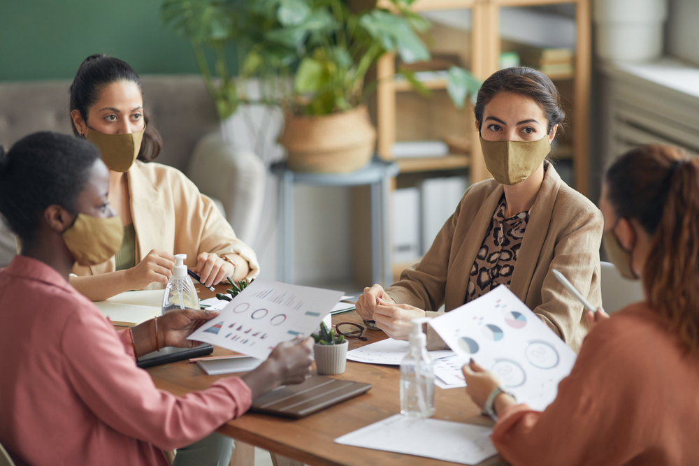 High angle view at multi-ethnic group of young businesswomen wearing masks during meeting in office, copy space